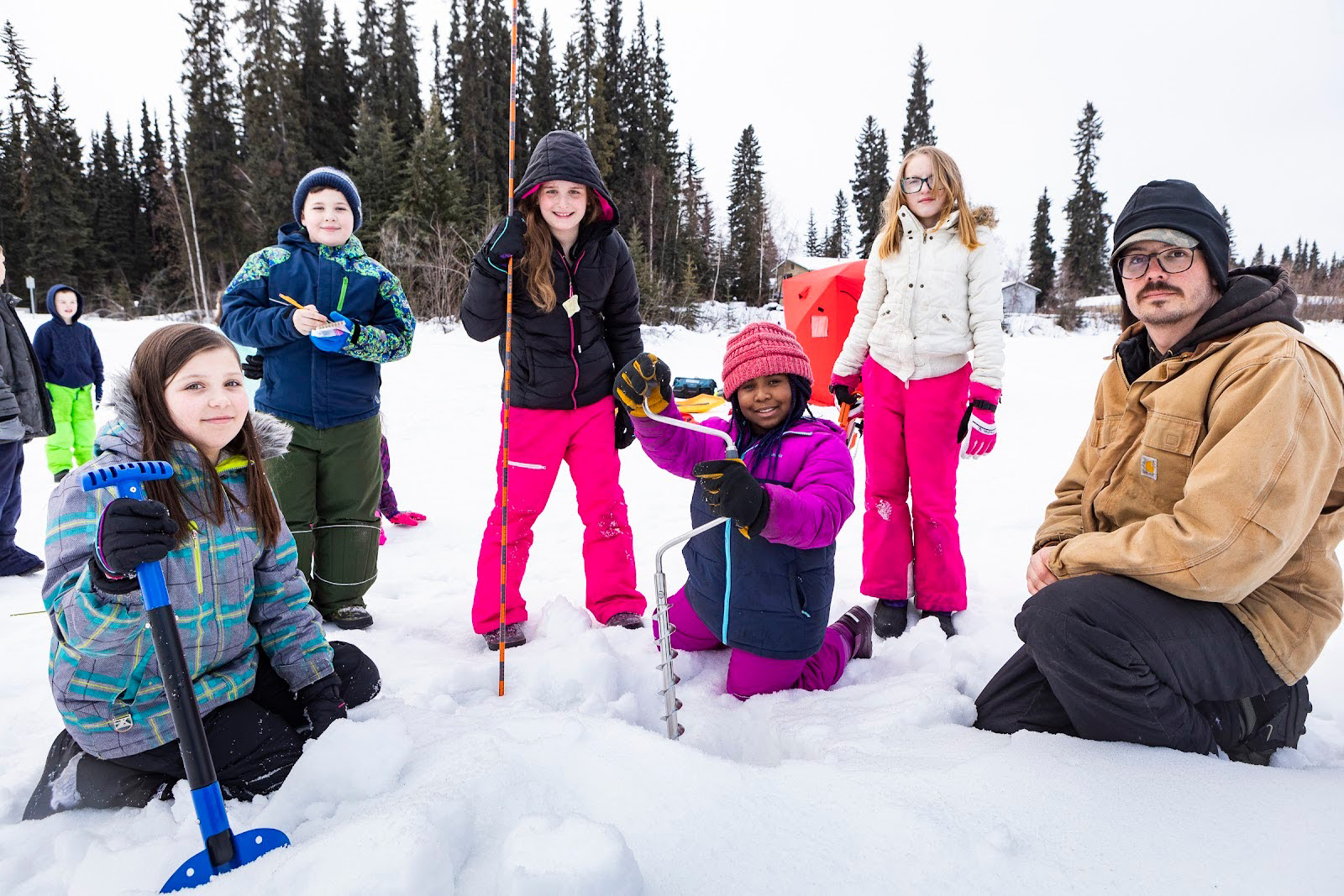 An adult and a group of children in colorful clothing drill a hole through the snow and ice.